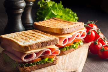 Close-up of two sandwiches with bacon, salami, prosciutto and fresh vegetables on rustic wooden cutting board. Club sandwich concept.