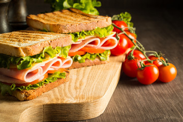 Close-up of two sandwiches with bacon, salami, prosciutto and fresh vegetables on rustic wooden cutting board. Club sandwich concept.