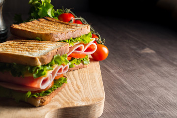 Close-up of two sandwiches with bacon, salami, prosciutto and fresh vegetables on rustic wooden cutting board. Club sandwich concept.