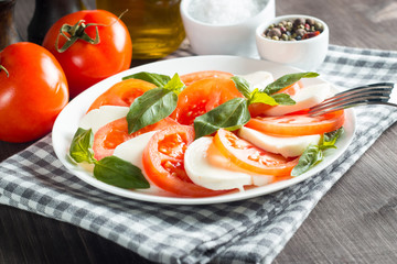 Photo of Caprese Salad with tomatoes, basil, mozzarella, olives and olive oil on wooden background. Italian traditional caprese salad ingredients. Mediterranean, organic and natural food concept.