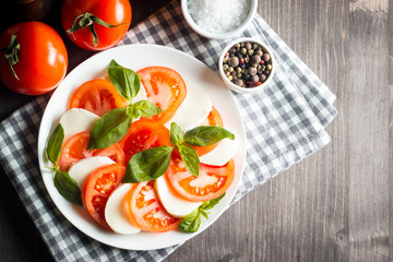Photo of Caprese Salad with tomatoes, basil, mozzarella, olives and olive oil on wooden background. Italian traditional caprese salad ingredients. Mediterranean, organic and natural food concept.