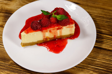 Piece of tasty New York cheesecake with raspberries and raspberry jam in a white plate on wooden table