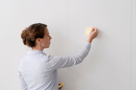 A business woman wearing a light blue shirt is pasting post it notes on a white wall.