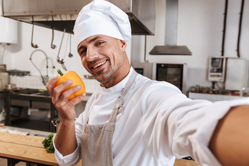 Photo of attractive male chief in apron taking selfie, while standing at kitchen in restaurant