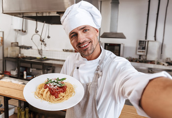 Image of happy man chief in apron taking selfie, while standing at kitchen in restaurant