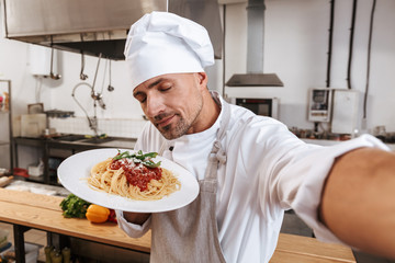 Image of professional man chief in apron taking selfie, while standing at kitchen in restaurant