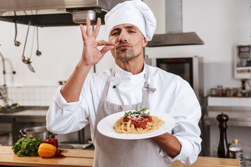 Photo of caucasian male chief in white uniform holding plate with meal, while cooking at kitchen in restaurant