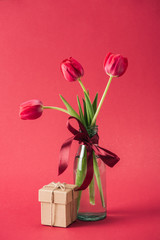 gift box and bouquet of red tulips with red satin bowl in glass vase on red background