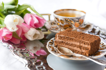 piece of chocolate cake on a plate, white and pink tulips, a mug of tea