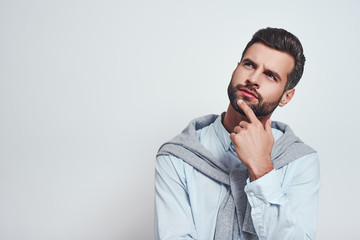 Thinking about the future...Close-up studio portrait of young bearded man keeping hand on chin, looking upwards and thinking. Grey background