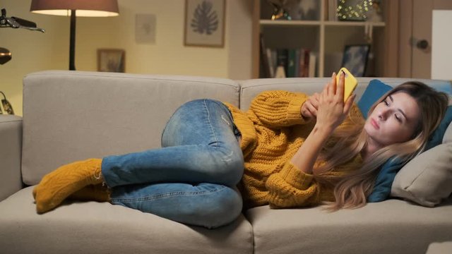 Tired woman texting on smartphone while lying on a sofa in the living room. Smiling blonde girl reading a message, browsing on internet and using app, having fun conversations chat with her mobile.