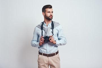 Confident photographer. Handsome young man in casual clothes is holding camera and looking away while standing on a grey background.