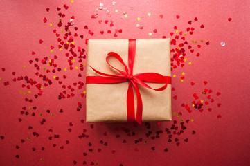 Gift box wrapped in brown colored craft paper and tied with red bow on dark red background with red heart shape confetti.