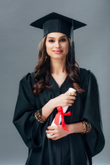 happy female indian student in academic gown and graduation hat holding diploma, isolated on grey
