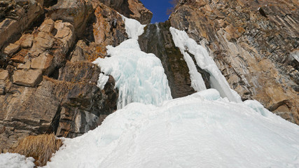 Fototapeta na wymiar Frozen waterfall among the rocks. The waterfall is freezing, huge icicles. Ice white and blue. Brown rocks and splashes of water. Winter waterfall. White snow and blue sky. Water runs down the ice.