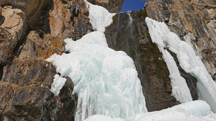 Fototapeta na wymiar Frozen waterfall among the rocks. The waterfall is freezing, huge icicles. Ice white and blue. Brown rocks and splashes of water. Winter waterfall. White snow and blue sky. Water runs down the ice.