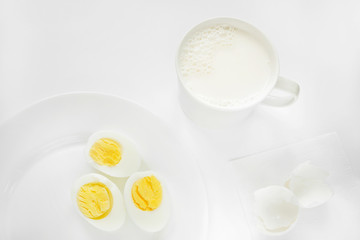 Very light photo with a predominance of white. On a white background there is a plate with sliced boiled eggs and a cup with milk.