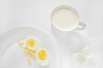 Very light photo with a predominance of white. On a white background there is a plate with sliced boiled eggs and a cup with milk.