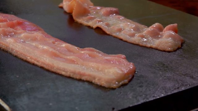 Closeup of a two bacon strips hissing and frying on the hot stone surface. Movement of the chamber along several pieces of bacon
