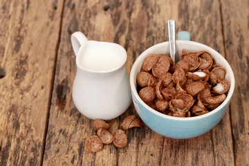 Chocolate cereal in a blue cup with tea spoon  and white jar of milk on the old wooden table, Close up