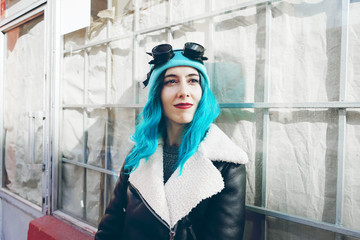 Portrait of a punk or gothic young woman with blue colored hair and wearing black steampunk glasses and blue wool cap in a urban outdoor street