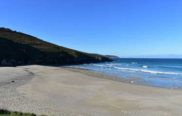 Beach with morning light. Golden sand, rocks and blue sea with waves and foam. Clear sky, sunny day. Galicia, Spain.