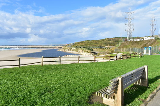 Beach with grass, wooden bench and fence. Industrial port and blue river, cloudy sky. Sabon, Galicia, Spain.