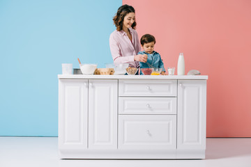 happy mother with adorable little son cooking together at white kitchen table on bicolor background