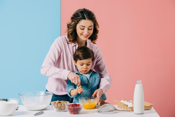 smiling mother helping little son in whipping eggs with balloon whisk on bicolor background