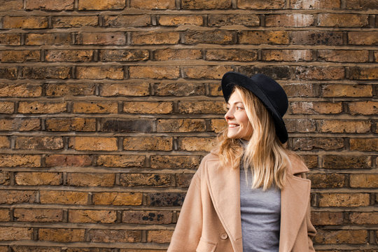 Portrait of beautiful smiling Caucasian woman standing by the brick wall wearing camel coat and a hat.