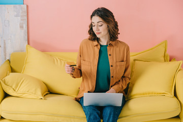 smiling attractive woman holding credit card while sitting on yellow sofa and using laptop