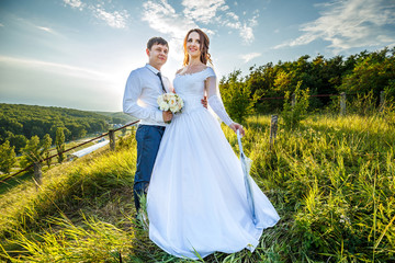 Fototapeta premium A young bridegroom gently hugs a beautiful bride with umbrella against the background of green hills and blue sky in sunny weather. Wedding photography.