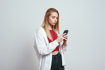 Always available. Young blonde woman in a casual wear using her smart phone while standing in studio on a grey background. Digital concept