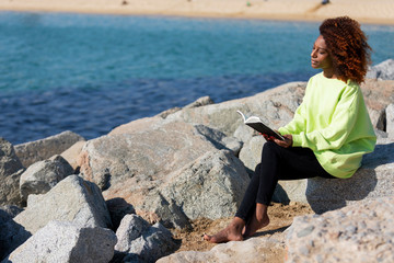 Side view of young curly afro woman sitting on a breakwater holding a book while reading outdoors