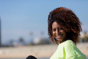 Side view of a beautiful curly afro woman sitting on breakwater rocks laughing while looking camera outdoors