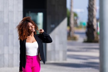 Front view of young beautiful curly woman wearing elegant clothes and handbag while standing in the street in sunny day