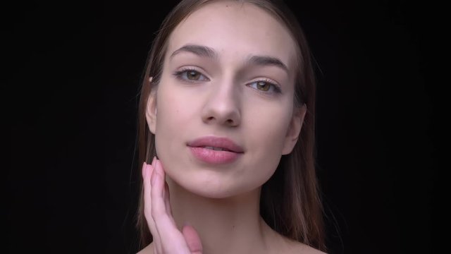 Young and slim caucasian girl with nude make-up watching into camera and touching her face with satisfaction on black background.