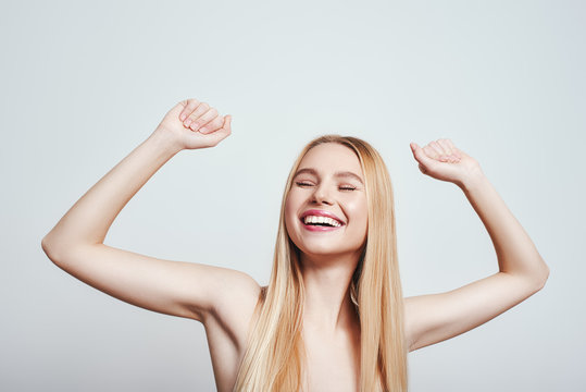 Yes! I did it! Happy young blonde woman with naked shoulders smiling with raised hands while standing on a grey background