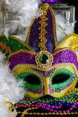 Jester mask propped up against glass block with feather boa and bright beads in the foreground.