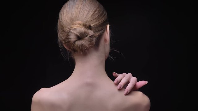 Rear portrait of young and slim caucasian girl touching softly her neck and back on black background.