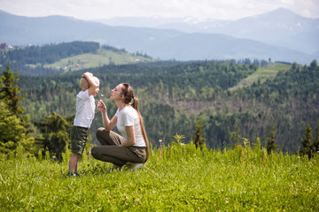 Mother and little son blow dandelion against the background of coniferous forest and mountains. Maternity and friendship