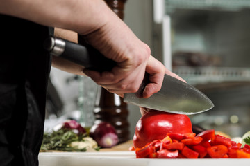 Male chef cuts vegetables for salad in a restaurant in a black apron. White cutting board, closeup of hands.