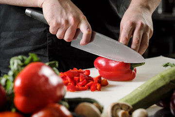 Male chef cuts vegetables for salad in a restaurant in a black apron. White cutting board, closeup of hands.