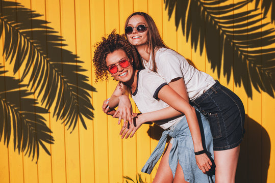 Candid shot of two lovely hipster women in glasses and denim clothes standing under the shadow of palm branches on a sunny summer day.