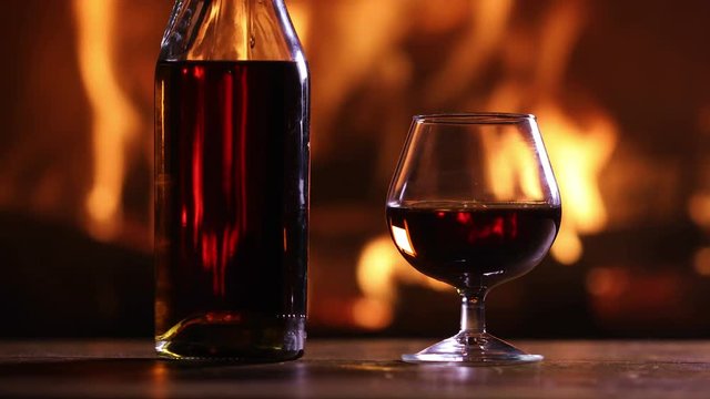 A bottle of brandy and a glass are on the table against the burning fireplace