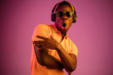 The young handsome happy hipster man listening music with headphones at black studio with neon lights. Disco, night club, hip hop style, positive emotions, face expression, dancing concept