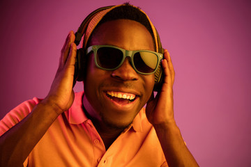 The young handsome happy hipster man listening music with headphones at black studio with neon lights. Disco, night club, hip hop style, positive emotions, face expression, dancing concept