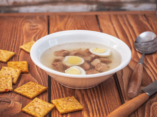 Lunch from broth with slices of boiled turkey, quail eggs in a deep white plate stand on a wooden table. A knife and spoon, a few crackers with herbs lie near the plate.