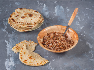 Buckwheat porridge in a deep plate and a tortilla torn into pieces stand on a wooden table. In the background, a plate with homemade cakes.