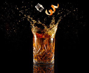 orange zest and ice falling into splashing cocktail isolated on black